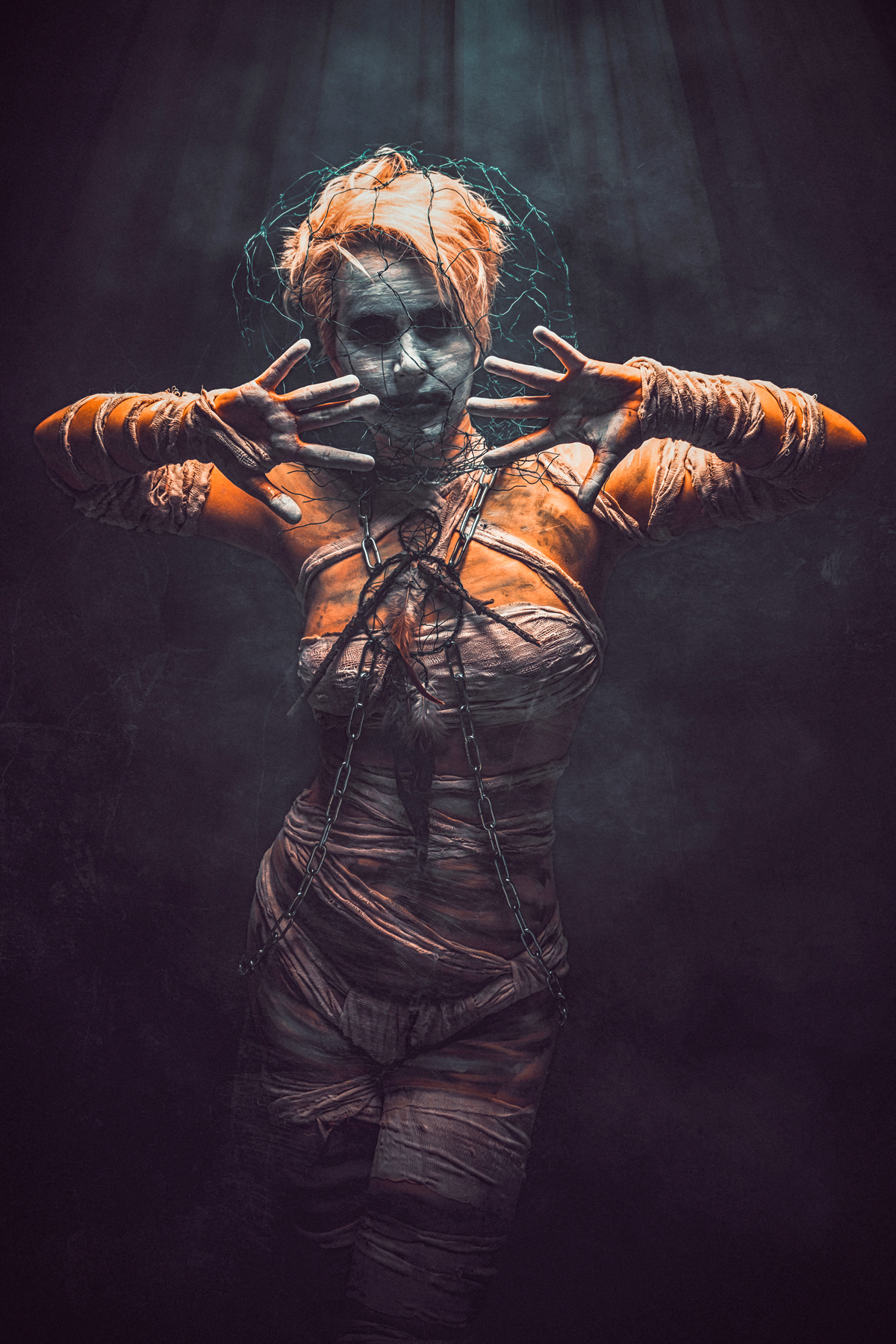 A cinematic dark art portrait of a woman wrapped in bandages and chains, featuring a wire mesh over her face and outstretched hands, emerging from a smoky, dark background with dramatic overhead lighting.