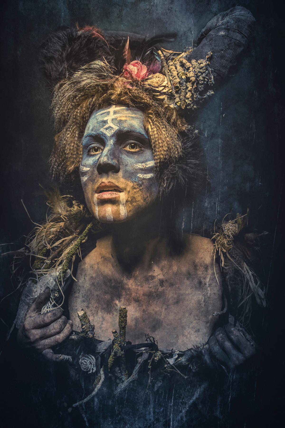 A tribal fine art portrait of a woman with blue and white ritual face paint, wearing a rustic headdress made of fur, feathers, and dried flowers, with mud-streaked skin and a dark, weathered background.