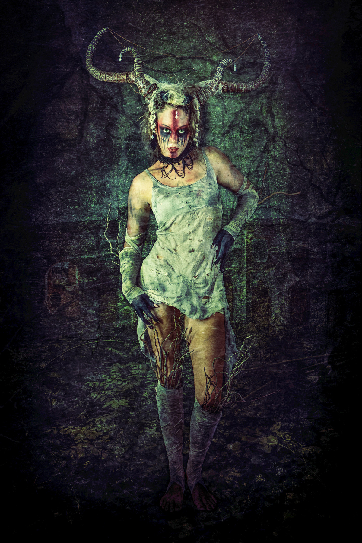 A full-body fine art portrait of a forest witch with large curved horns, wearing a tattered white dress and leg wraps with dry branches protruding. The figure has dark ritual face paint and black-stained hands, set against a dark, overgrown forest background.