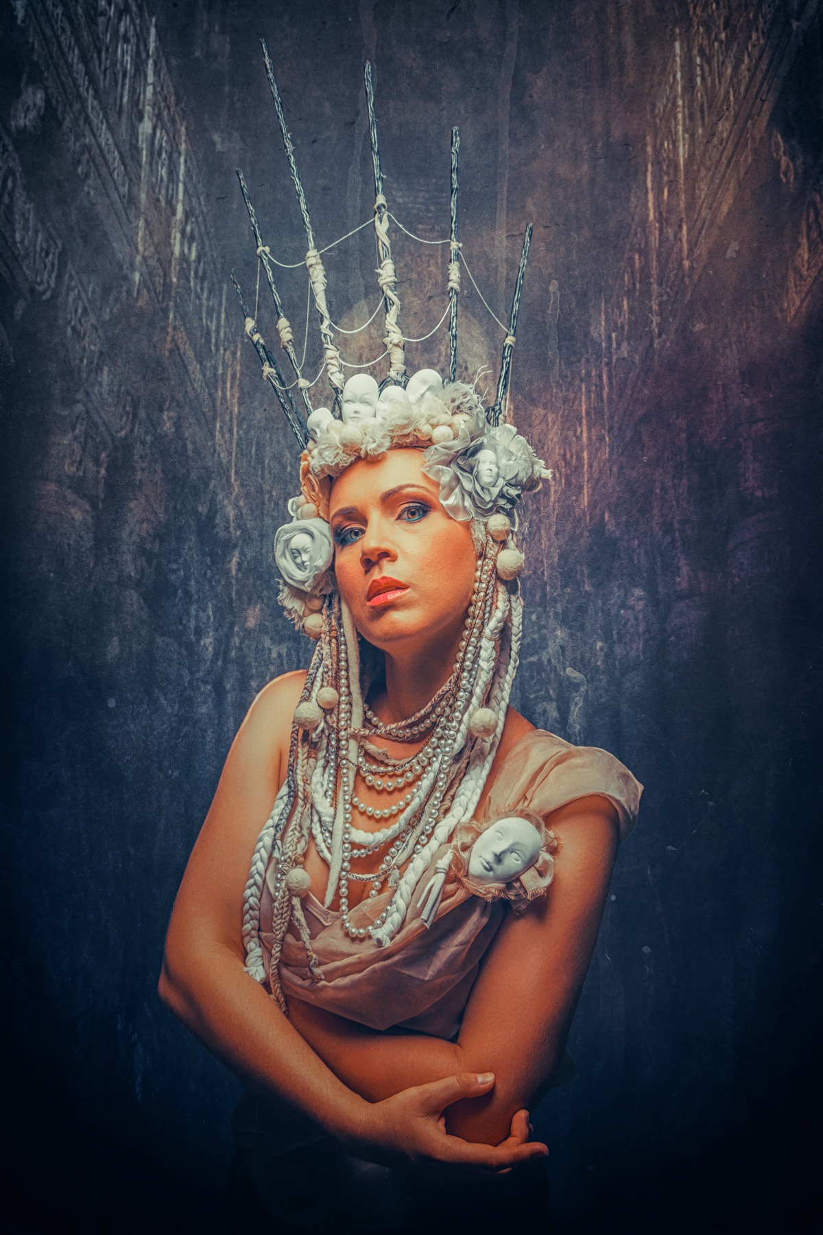 A surreal fine art portrait of a woman wearing an elaborate crown made of spikes, spiderwebs, and small doll faces, adorned with pearls, braided white hair, and a miniature porcelain mask on her shoulder.