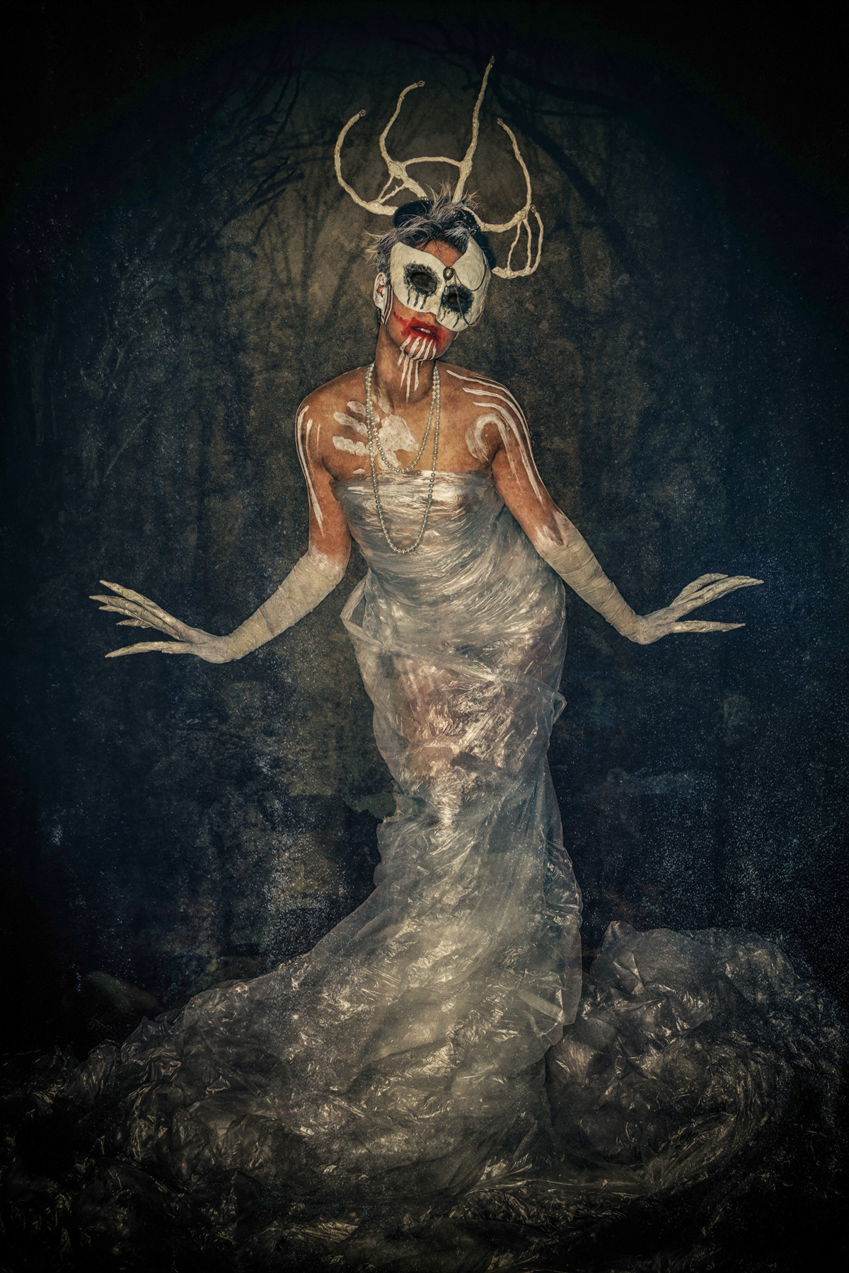 A full-body fine art portrait of a spectral queen wrapped in translucent plastic film, wearing an antler-like headdress and a white mask with bleeding red accents, featuring white ritual body paint and long, claw-like fingers
