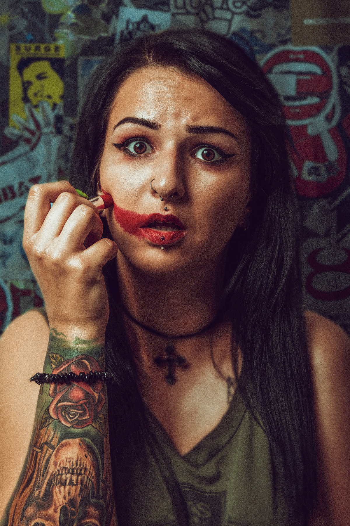 A gritty urban portrait of a woman with facial piercings and a large forearm tattoo, applying red lipstick across her cheek in a wide, smeared stroke, set against a background of stickers and graffiti.